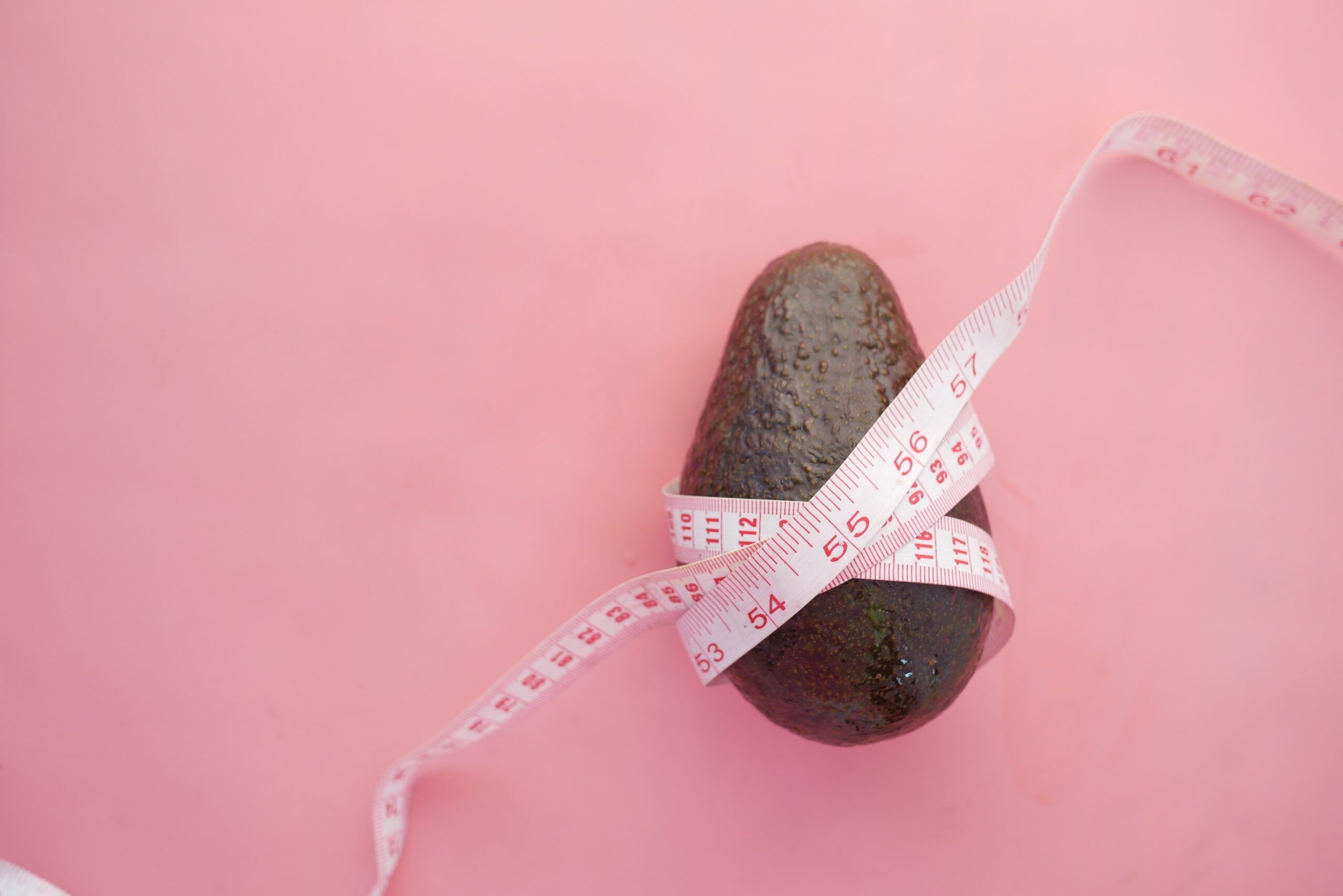 Photograph of an avocado resting on a pink tabletop with a measuring tape around it.