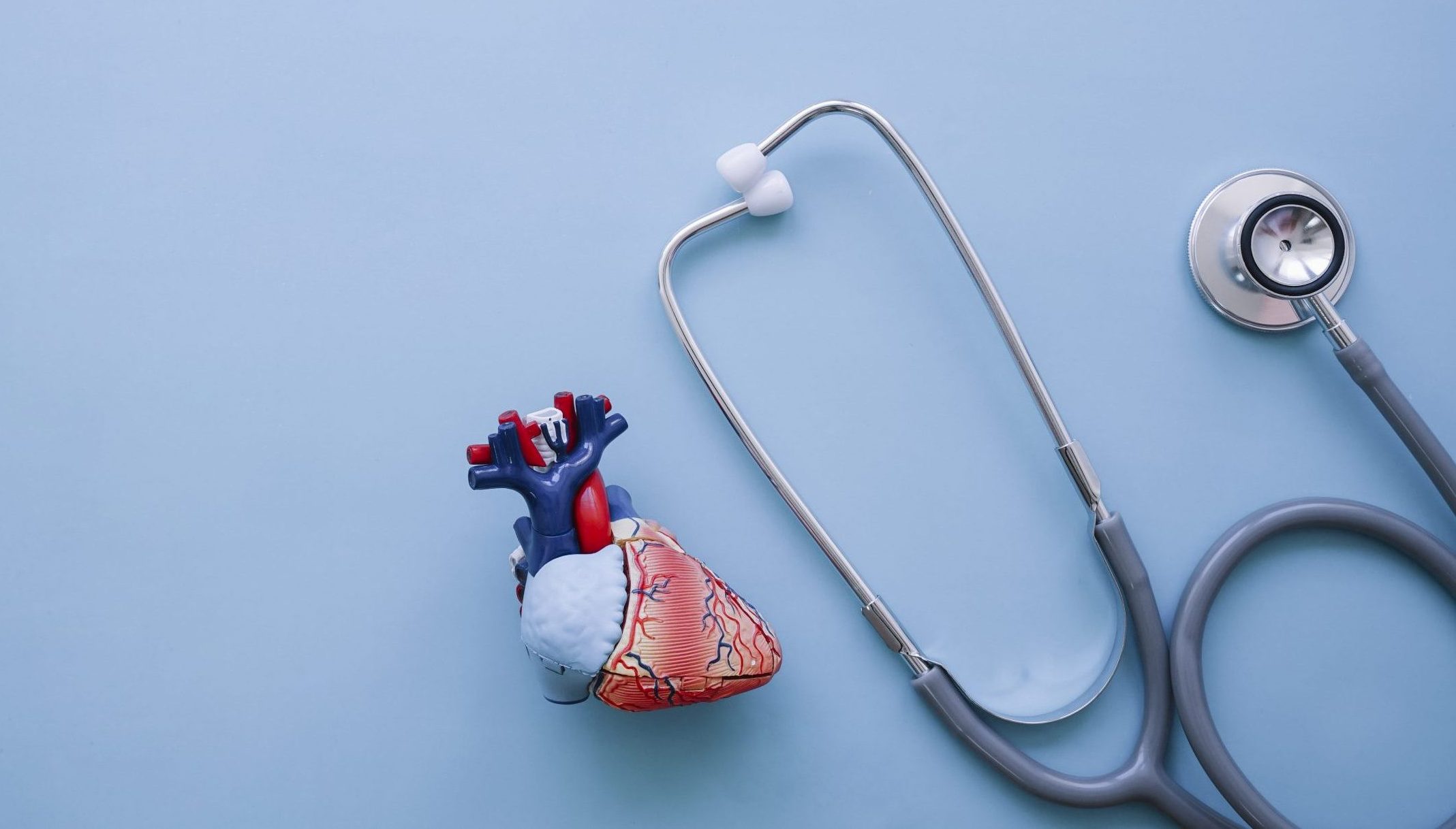 Image of a stethoscope next to a model heart on a blue background.